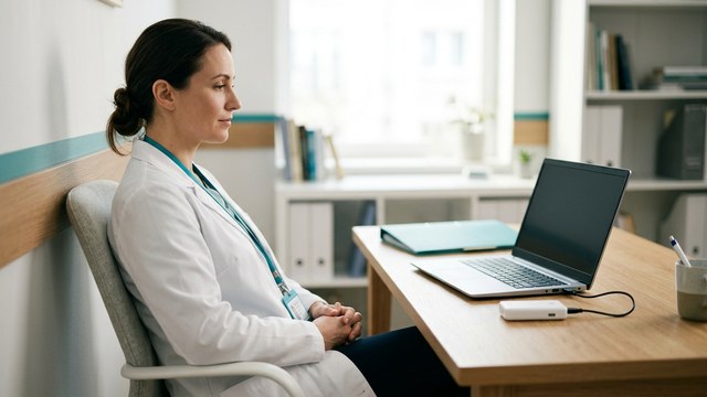 Lekarka w gabinecie przy laptopie z podłączonym niewielkim urządzeniem, dłonie z dala od klawiatury